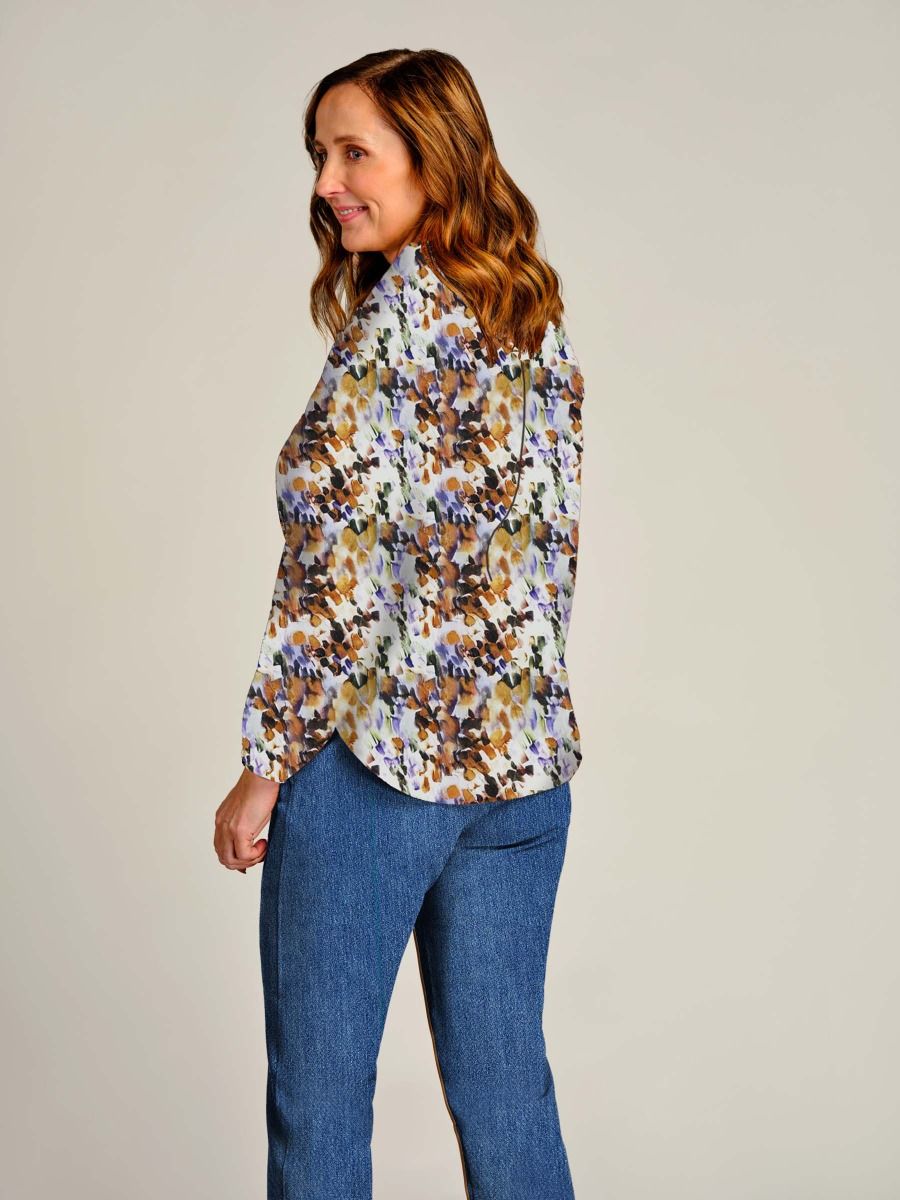 A woman wearing a long-sleeve button-up blouse with a dense abstract floral print in brown, blue, purple, and white, paired with blue jeans, photographed against a solid light gray studio background.