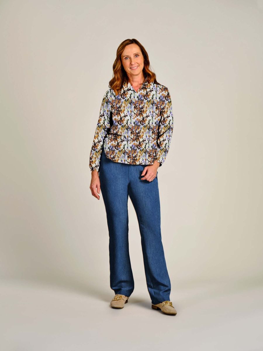 A woman stands against a plain light beige studio background wearing a long-sleeve button-up shirt with a dense multicolor floral print in blue, yellow, white, and brown tones, paired with blue jeans and tan shoes.