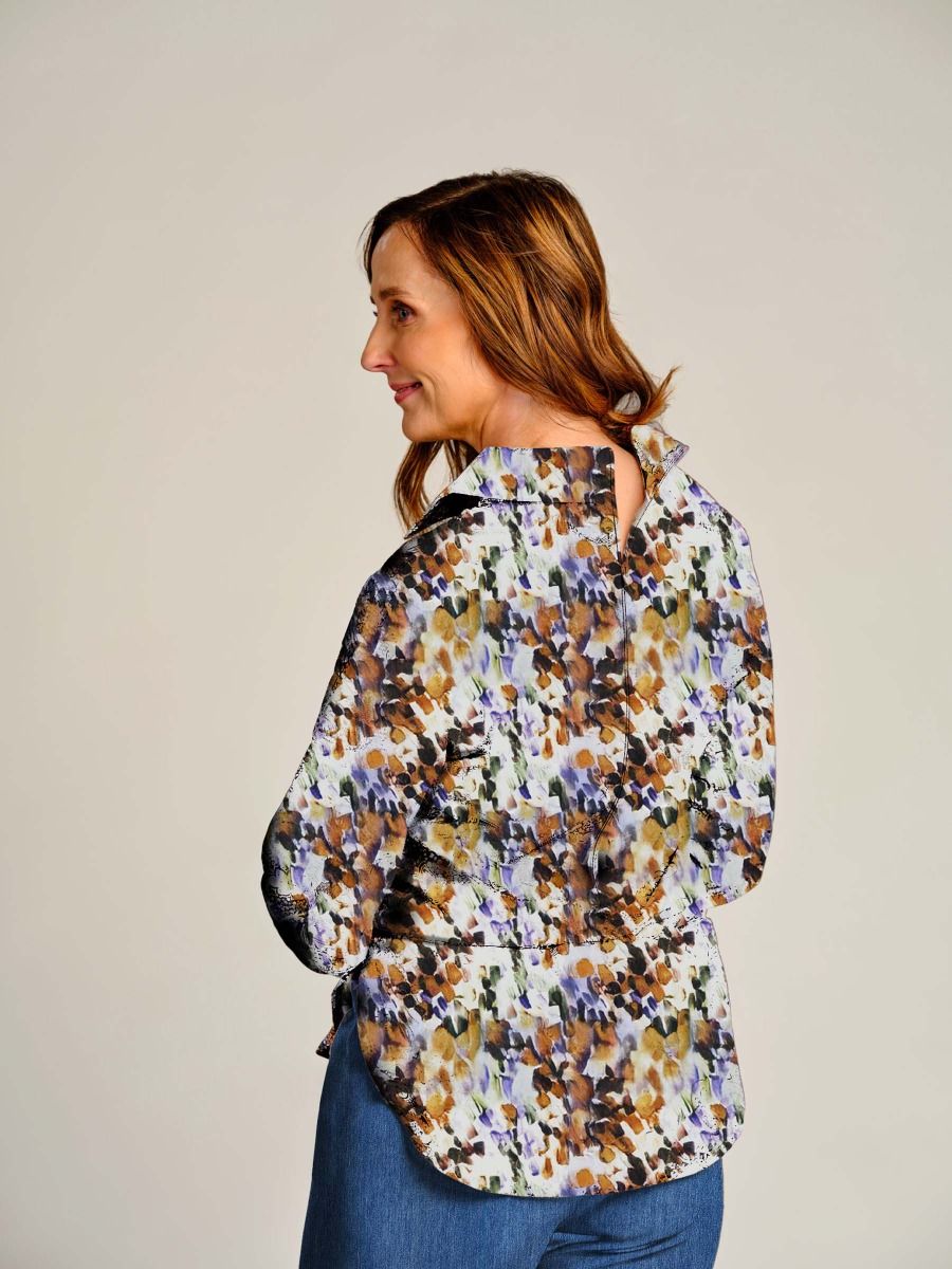 A woman shown from the back wearing a long-sleeve button-up shirt with an abstract floral pattern in brown, purple, and orange on a white background, paired with blue jeans, against a plain light gray studio background.