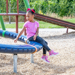 A young girl with her hair in two buns sits on a blue playground merry-go-round. She is wearing a pink t-shirt, dark gray leggings, and pink sneakers, set against a background of a playground with wood chips, green grass, and trees.