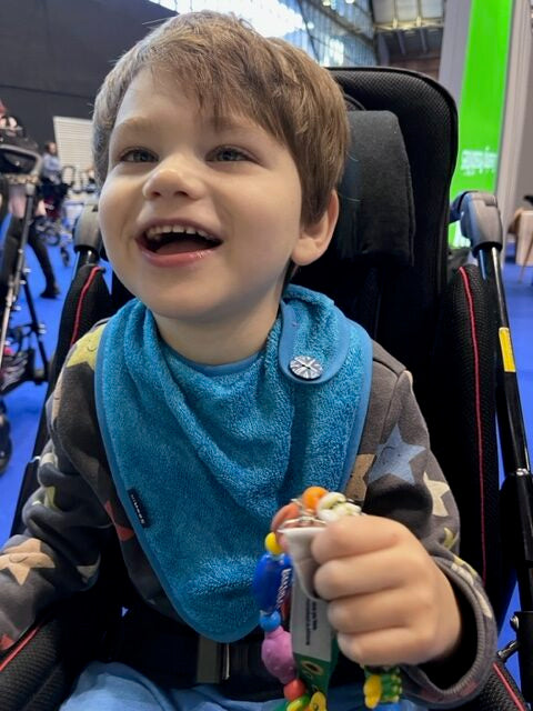 A young boy smiling in a black stroller at an indoor event, wearing a bright blue bib with a small white circular logo.