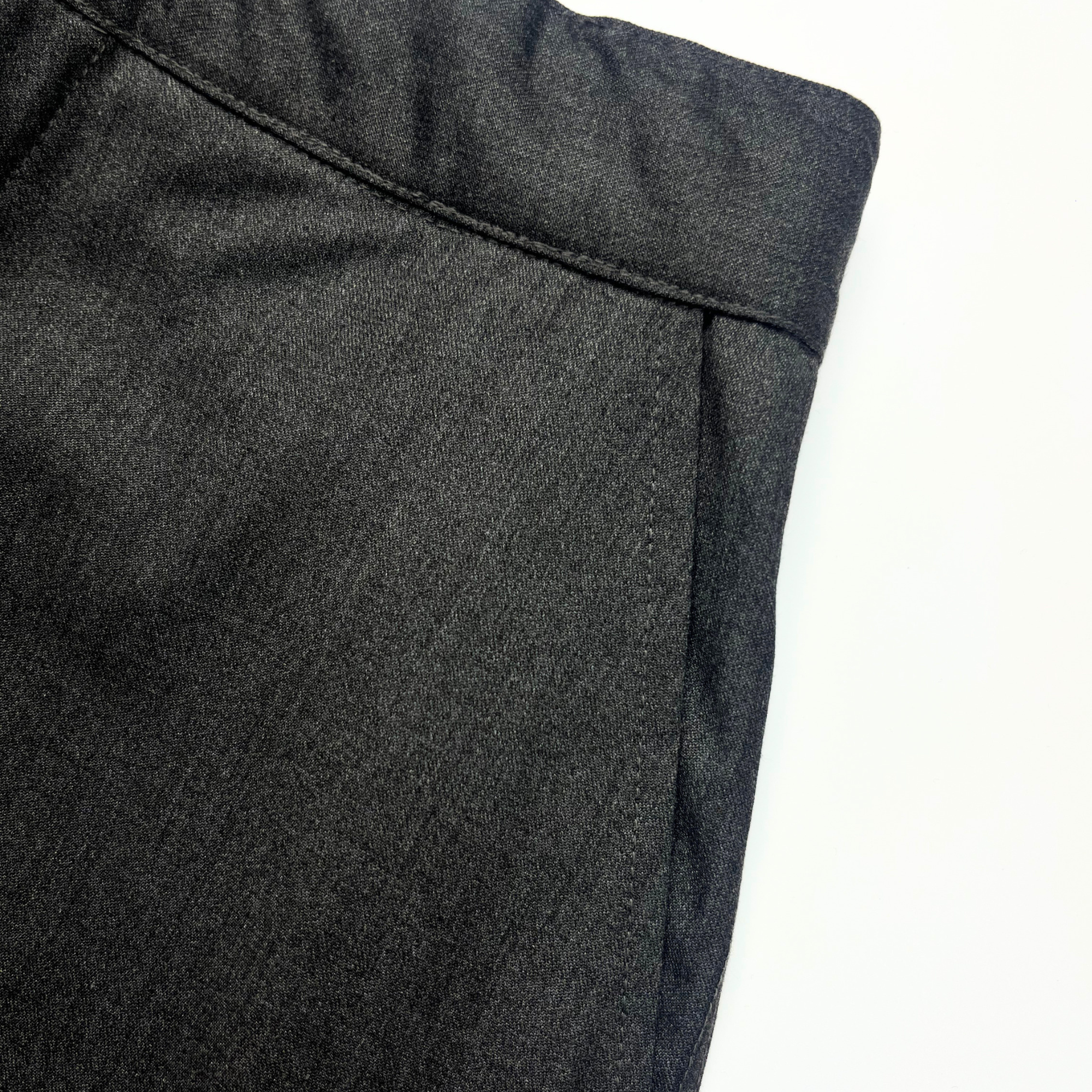 Close-up of dark charcoal gray textured fabric, likely trousers or a jacket, showing a waistband seam and side seam against a plain white background.
