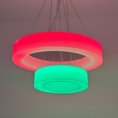 A modern two-ring LED pendant light hanging from chains against a plain ceiling. The top ring is illuminated in bright pink-red, and the bottom ring is illuminated in bright green.