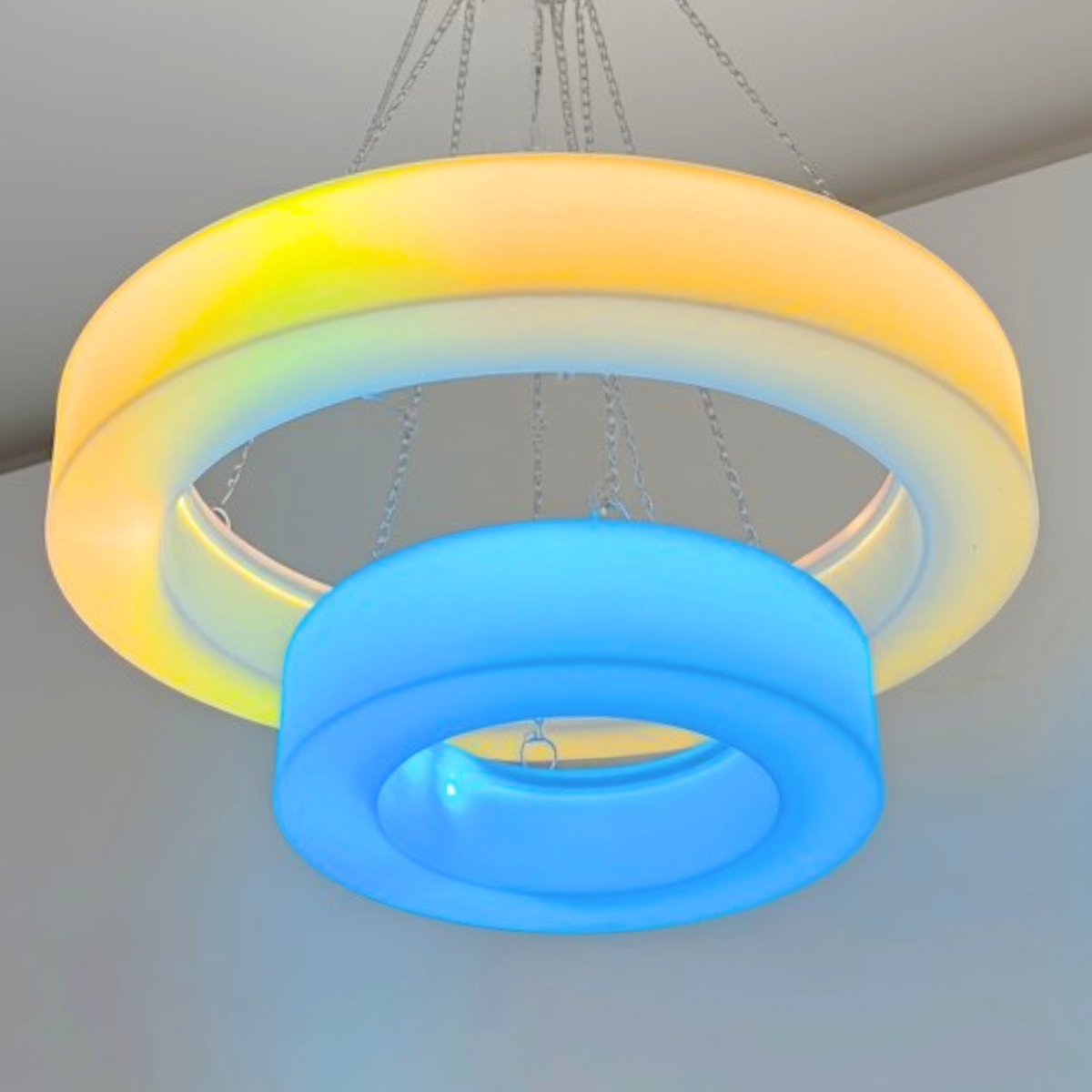 A modern two-ring LED ceiling light fixture suspended by chains. The larger outer ring is illuminated in a warm yellow-to-white gradient, while the smaller inner ring in front glows in a cool blue light.