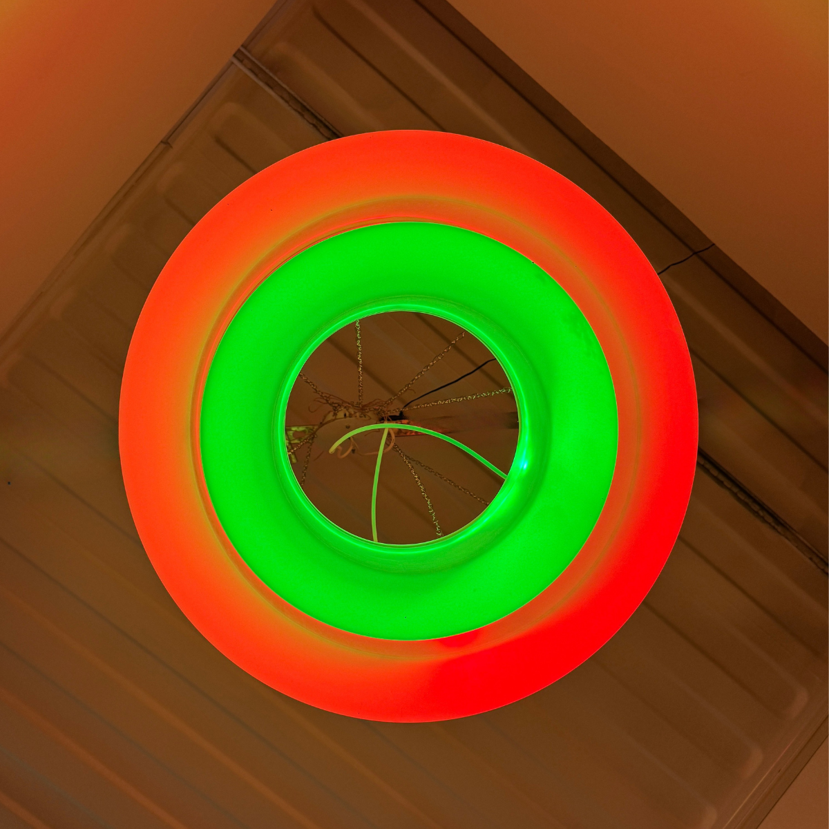 A circular LED light fixture with concentric rings glowing in red and green, mounted on a beige ribbed ceiling.