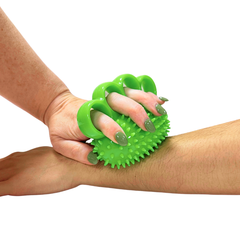 A bright green claw-shaped arnica massage tool with spiky edges being held against a person's forearm, shown on a plain white background.