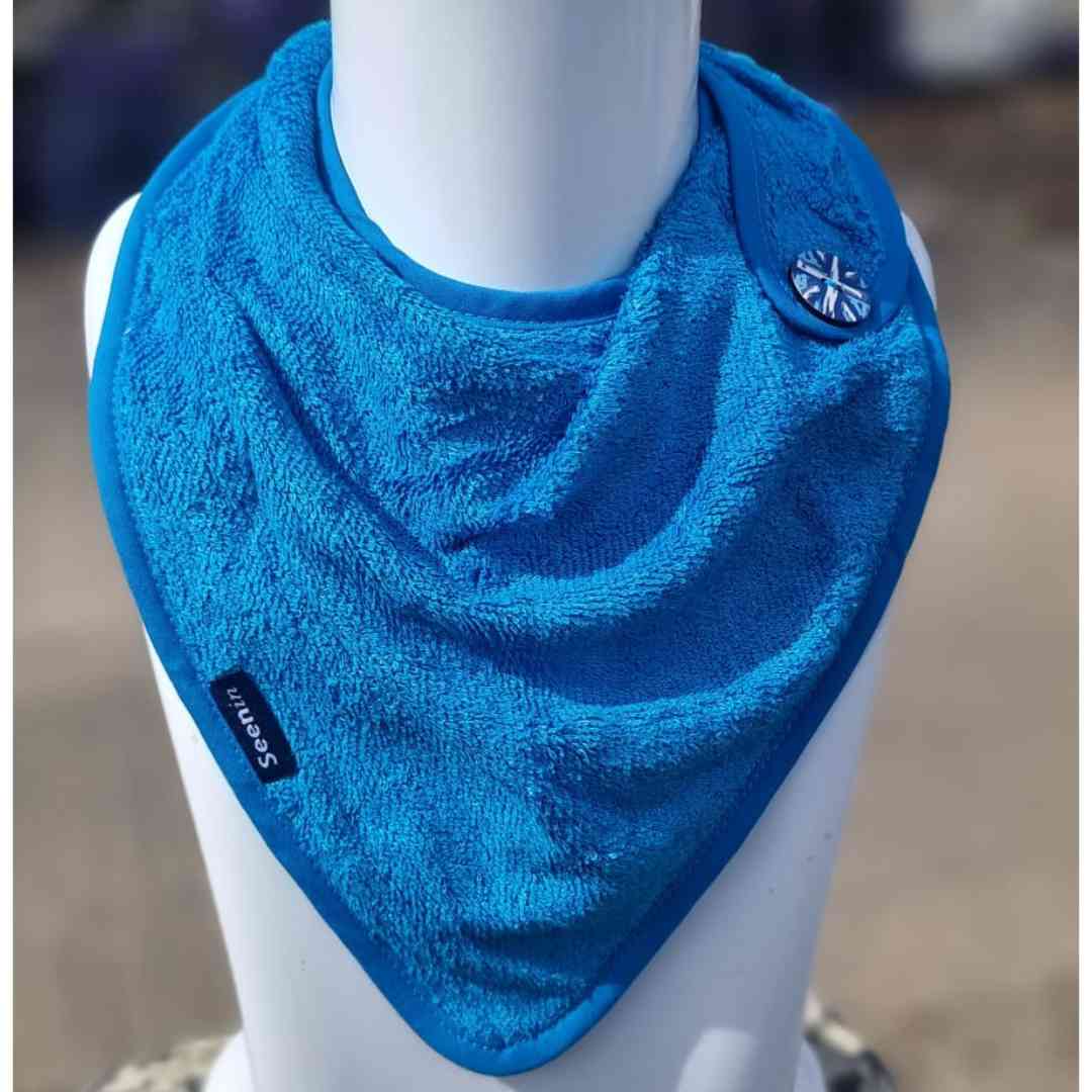 A bright blue triangular bib or neck gaiter made of soft terry cloth, displayed on a white mannequin torso. The bib features a small black brand tag on the lower left corner and a silver-toned decorative emblem on the upper right corner.